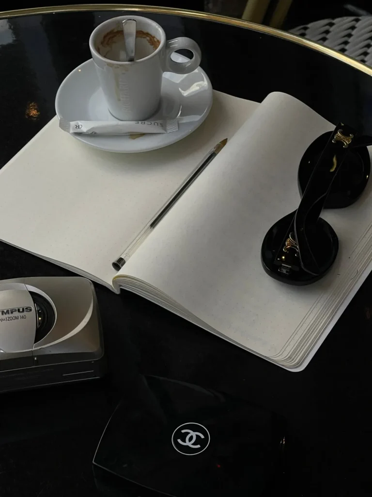 Chic café scene with coffee, notebook, pen, and sunglasses on a table.