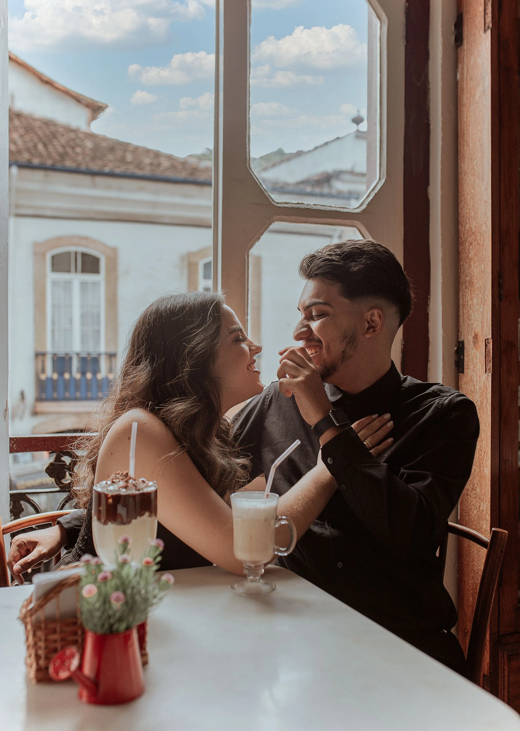 Two people smiling at each other across a cafe table by an open window