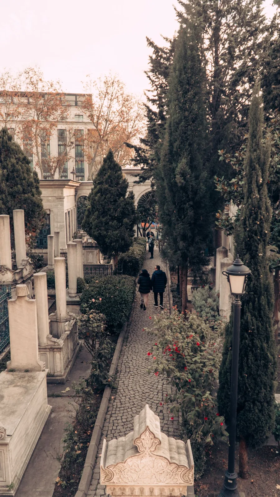 A couple wanders through a serene park with cobblestones and tall trees on a quiet day.