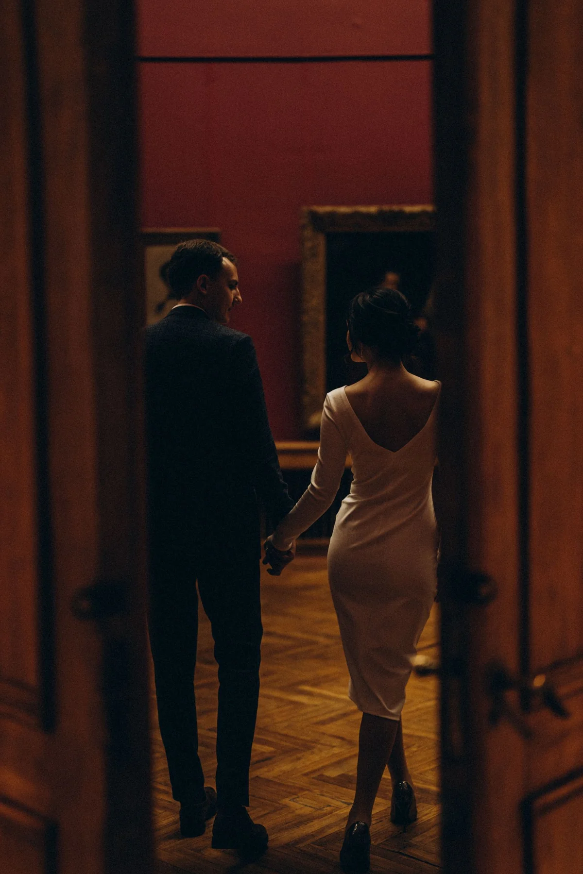 A couple in formal attire walks hand in hand inside a dimly lit art gallery, creating a romantic atmosphere.