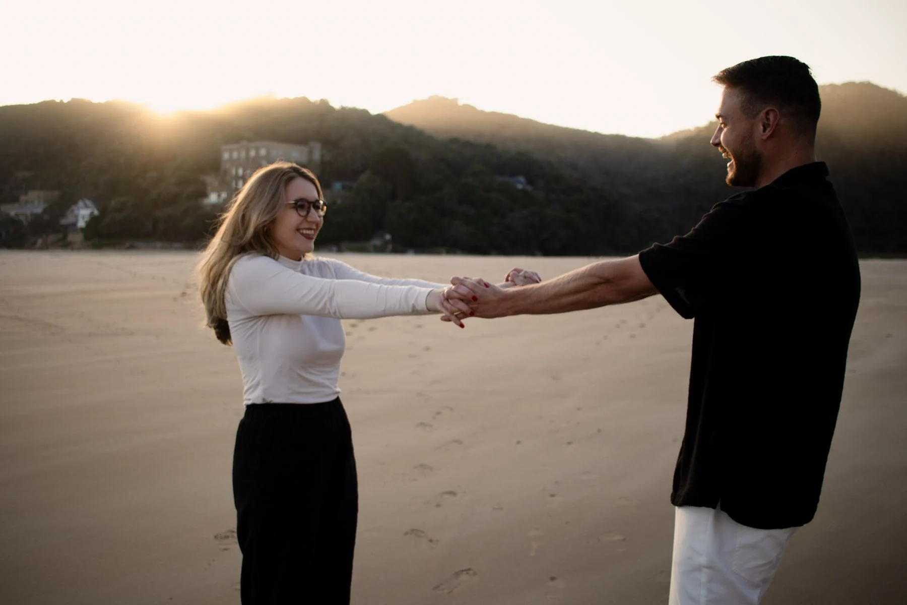 A couple holding hands on a sandy beach during sunset, displaying love and joy.
