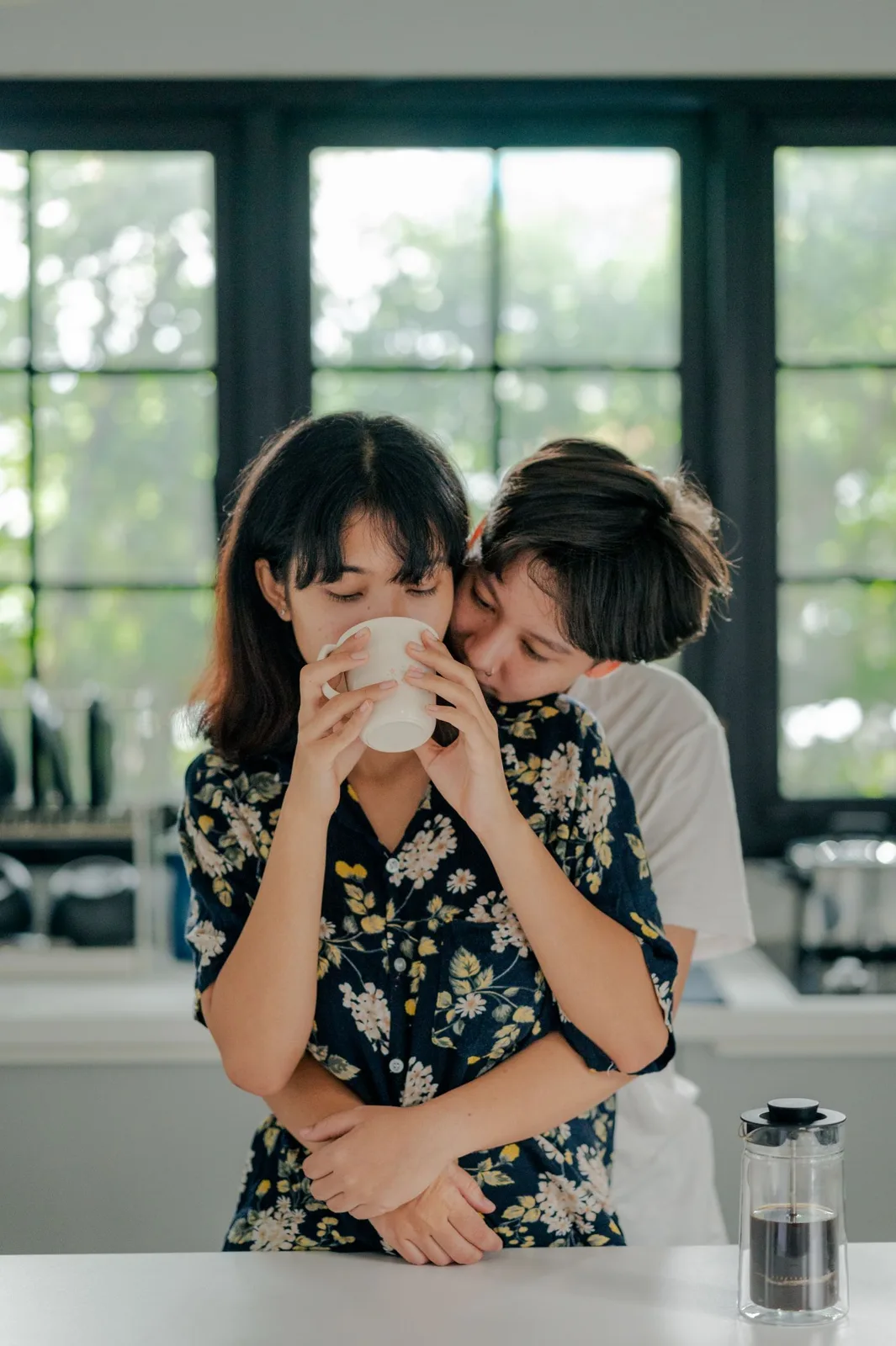 A couple shares a tender moment with coffee in a bright kitchen by a window.
